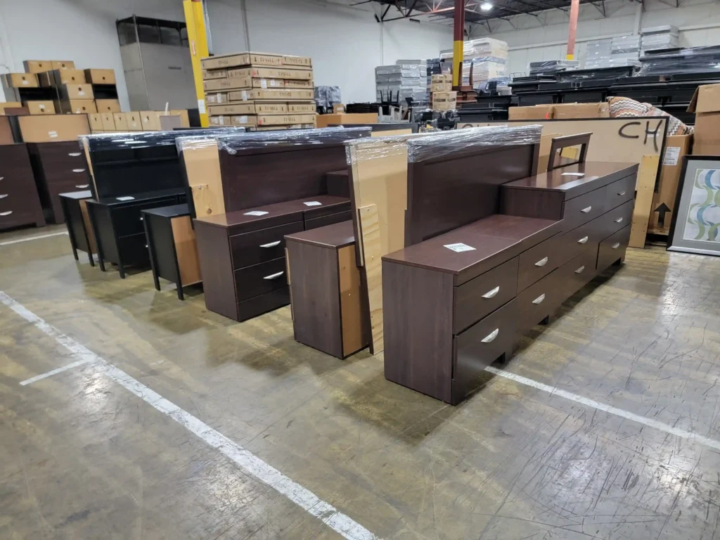 “Warehouse display of bedroom furniture for sale, including dresser, mirror, nightstands, TV stand, and boxed items with throw pillows.”
