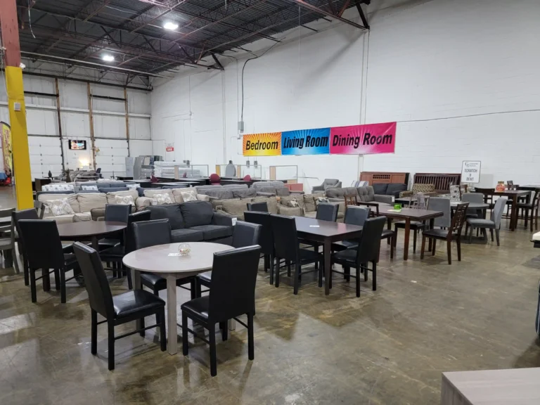 “Dining tables and chairs displayed in a warehouse, with sofas, loveseats, and signs for bedroom, living, and dining rooms in the background.”