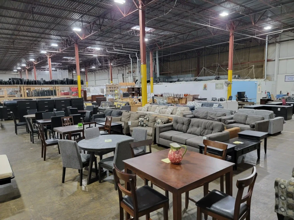 “Warehouse display of dining tables and chairs, with living room furniture, coffee tables, and bedroom furniture behind.”