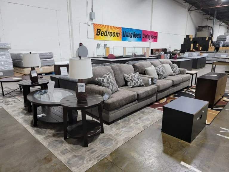 “Gray sofa and loveseat with throw pillows, coffee tables and TV stands on warehouse display, more furniture in the background.”