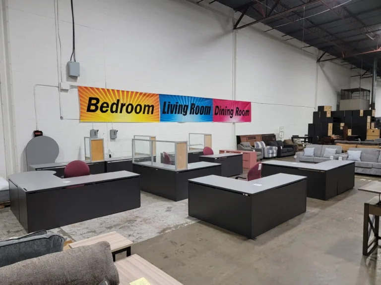 “Warehouse display of L-shaped desk and office chairs, with living room furniture and bedroom/living/dining room signs behind.”