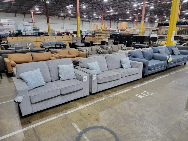“Warehouse display of gray, blue, caramel, and brown sofa and loveseat sets with bedroom and dining furniture in the back.”
