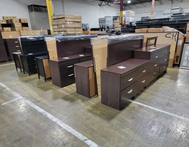 “Warehouse display of bedroom furniture for sale, including dresser, mirror, nightstands, TV stand, and boxed items with throw pillows.”