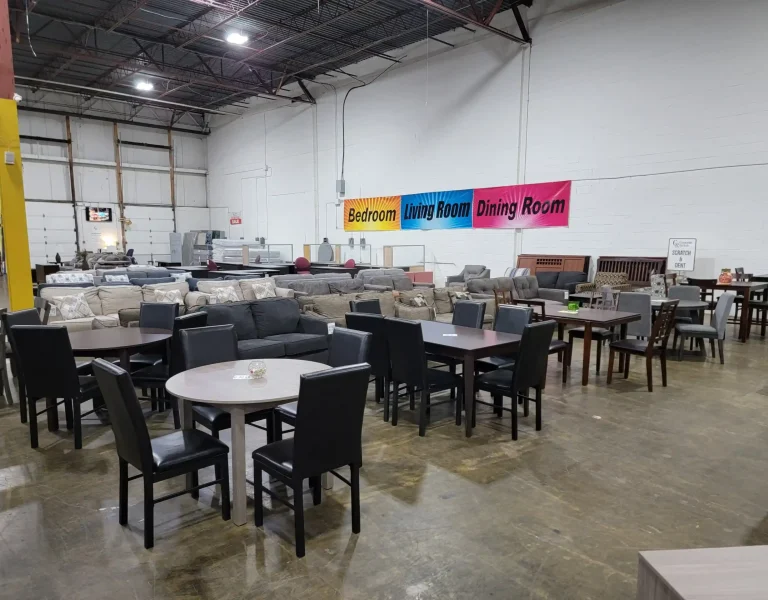 “Dining tables and chairs displayed in a warehouse, with sofas, loveseats, and signs for bedroom, living, and dining rooms in the background.”
